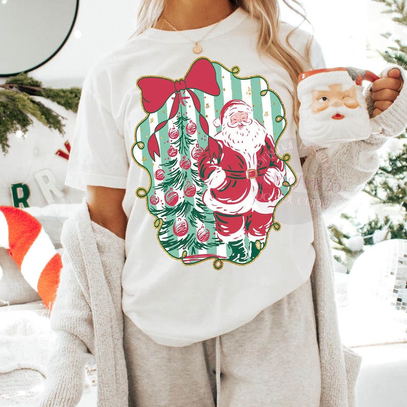 May include: White t-shirt featuring a Christmas design. The graphic includes Santa Claus in a red suit, a Christmas tree with red ornaments, and a large red bow. The design is framed with a gold border.