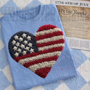 Peut inclure: T-shirt bleu clair avec un motif en forme de cœur représentant le drapeau américain. Le drapeau est fait de matière texturée rouge, blanche et bleue. Le texte "1776 4th of July" et "We the People" est visible en arrière-plan.