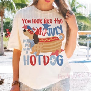 May include: Cream-colored t-shirt with a graphic of a hot dog dressed as a dog, with the text "You look like the 4th of July" and "makes me want a HOTDOG". The design incorporates red, white, and blue colors.