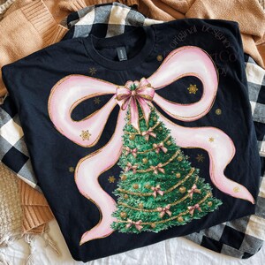 Pu&ograve; includere: T-shirt nera con un motivo a forma di albero di Natale. Il design include un grande fiocco rosa, un albero di Natale verde decorato con fiocchi rosa e ornamenti dorati e fiocchi di neve dorati. Il testo "Original Designer Art Co" &egrave; visibile.