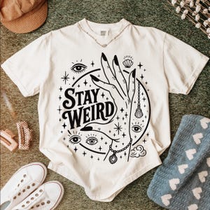May include: Cream-colored t-shirt with a black graphic design featuring a hand, eyes, stars, and the words "Stay Weird." The design includes a necklace and a seashell. A silver necklace is around the neckline.