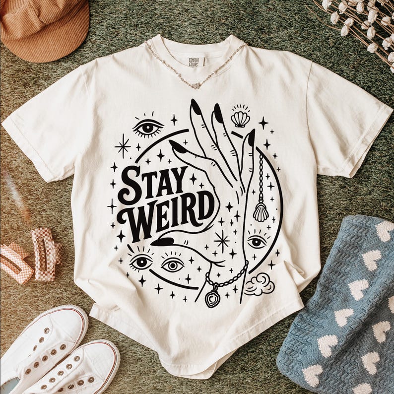 May include: Cream-colored t-shirt with a black graphic design featuring a hand, eyes, stars, and the words "Stay Weird." The design includes a necklace and a seashell. A silver necklace is around the neckline.