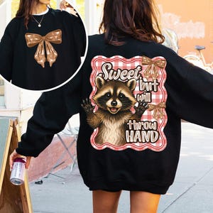 May include: Black sweatshirt featuring a brown bow on the front and a raccoon graphic on the back. The raccoon graphic is set against a red and white checkered background and includes the text "Sweet but will throw hand."