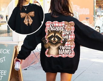 May include: Black sweatshirt featuring a brown bow on the front and a raccoon graphic on the back. The raccoon graphic is set against a red and white checkered background and includes the text "Sweet but will throw hand."