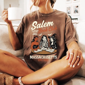May include: Brown t-shirt with the text "Salem Book Club" and an illustration of three witches reading books. The shirt also includes the text "Massachusetts Est. 1692".