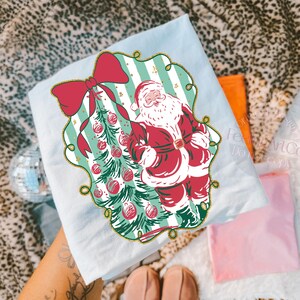 May include: Light blue t-shirt featuring a vintage illustration of Santa Claus next to a Christmas tree adorned with red ornaments. The design is framed by a red bow and a gold border. The shirt is folded.