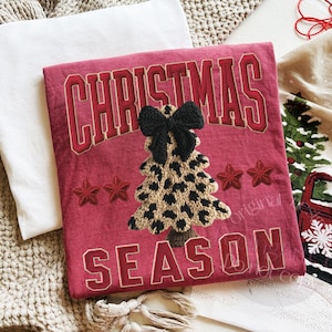 May include: A red Christmas-themed decorative pillow with the words "CHRISTMAS SEASON" in a collegiate font. The pillow features an embroidered leopard print Christmas tree with a black bow and red stars.