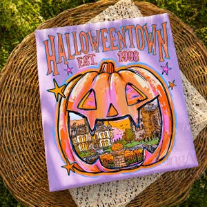 May include: A light purple t-shirt featuring a large orange jack-o'-lantern graphic with a detailed illustration of a house inside. The text HALLOWEENTOWN EST. 1998 is printed in orange above the pumpkin, accented by small pink and orange stars.