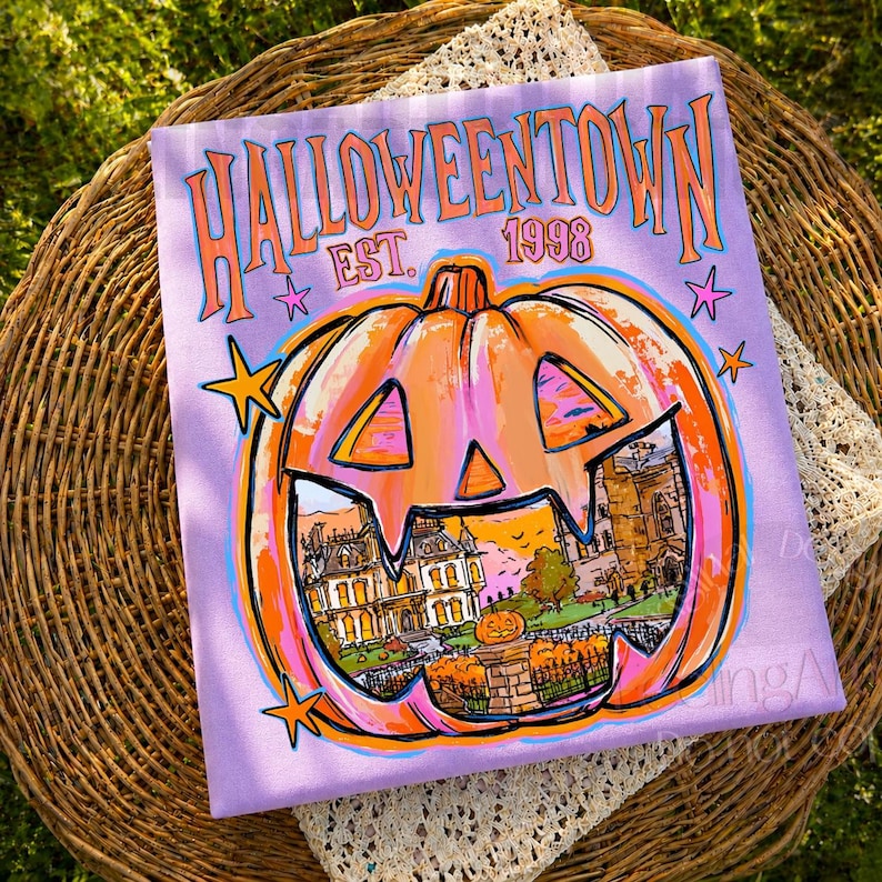 May include: A light purple t-shirt featuring a large orange jack-o'-lantern graphic with a detailed illustration of a house inside. The text HALLOWEENTOWN EST. 1998 is printed in orange above the pumpkin, accented by small pink and orange stars.