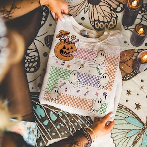 May include: A beige Halloween-themed sweatshirt featuring a jack-o'-lantern filled with candy and cartoon ghosts. The design includes colorful checkered stripes and scattered candies and lollipops. The text "Feeling Spooky" is visible.