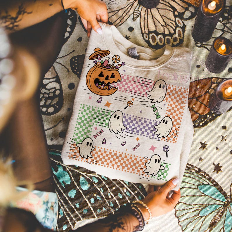 May include: A beige Halloween-themed sweatshirt featuring a jack-o'-lantern filled with candy and cartoon ghosts. The design includes colorful checkered stripes and scattered candies and lollipops. The text "Feeling Spooky" is visible.