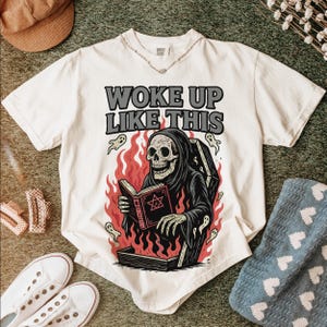 May include: White t-shirt featuring the text "WOKE UP LIKE THIS" and a graphic of a skeleton in a black robe reading a book, set against flames. Ghost illustrations are also included.