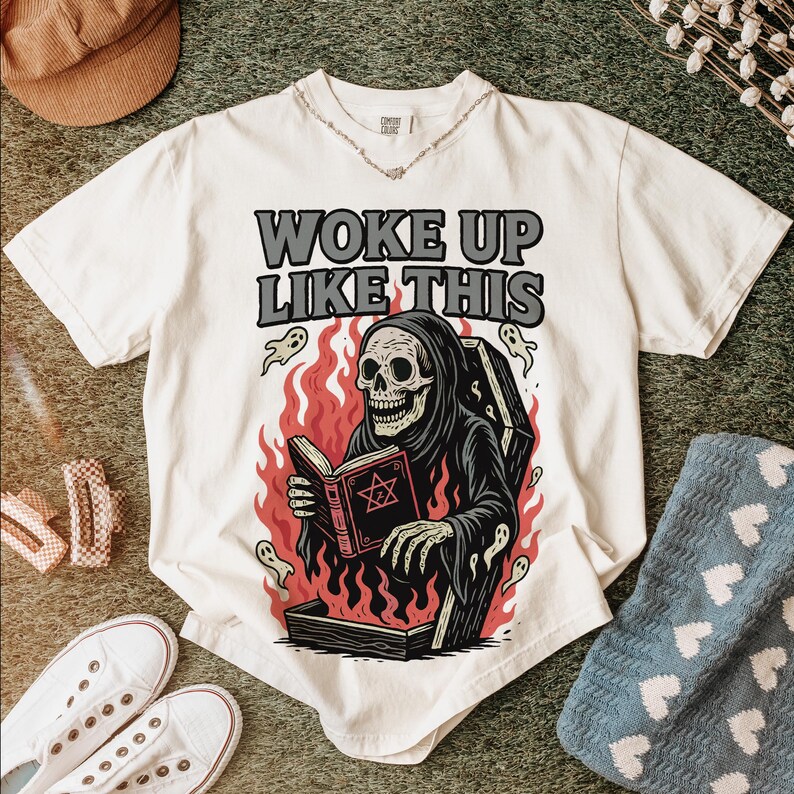 May include: White t-shirt featuring the text "WOKE UP LIKE THIS" and a graphic of a skeleton in a black robe reading a book, set against flames. Ghost illustrations are also included.