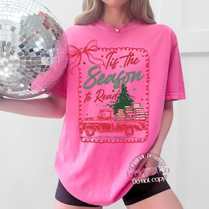 May include: A vibrant pink t-shirt with the text "Tis the Season to Read." The graphic shows a red truck laden with books and a Christmas tree. A red bow and decorative border frame the text.