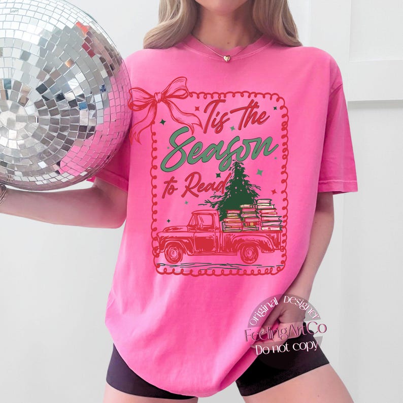 May include: A vibrant pink t-shirt with the text "Tis the Season to Read." The graphic shows a red truck laden with books and a Christmas tree. A red bow and decorative border frame the text.