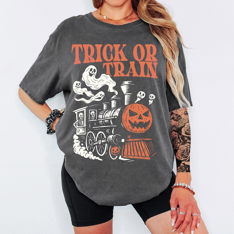 May include: Dark grey t-shirt with the words "TRICK OR TRAIN" in orange. The graphic features a Halloween-themed train with a jack-o'-lantern, ghosts, and skulls. A fun and festive design for the spooky season.