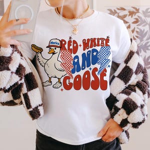 May include: White t-shirt featuring a cartoon goose wearing a baseball cap with "Merica" on it, holding a hot dog. The shirt has the text "Red White and Goose" in red, white, and blue. A casual style t-shirt.