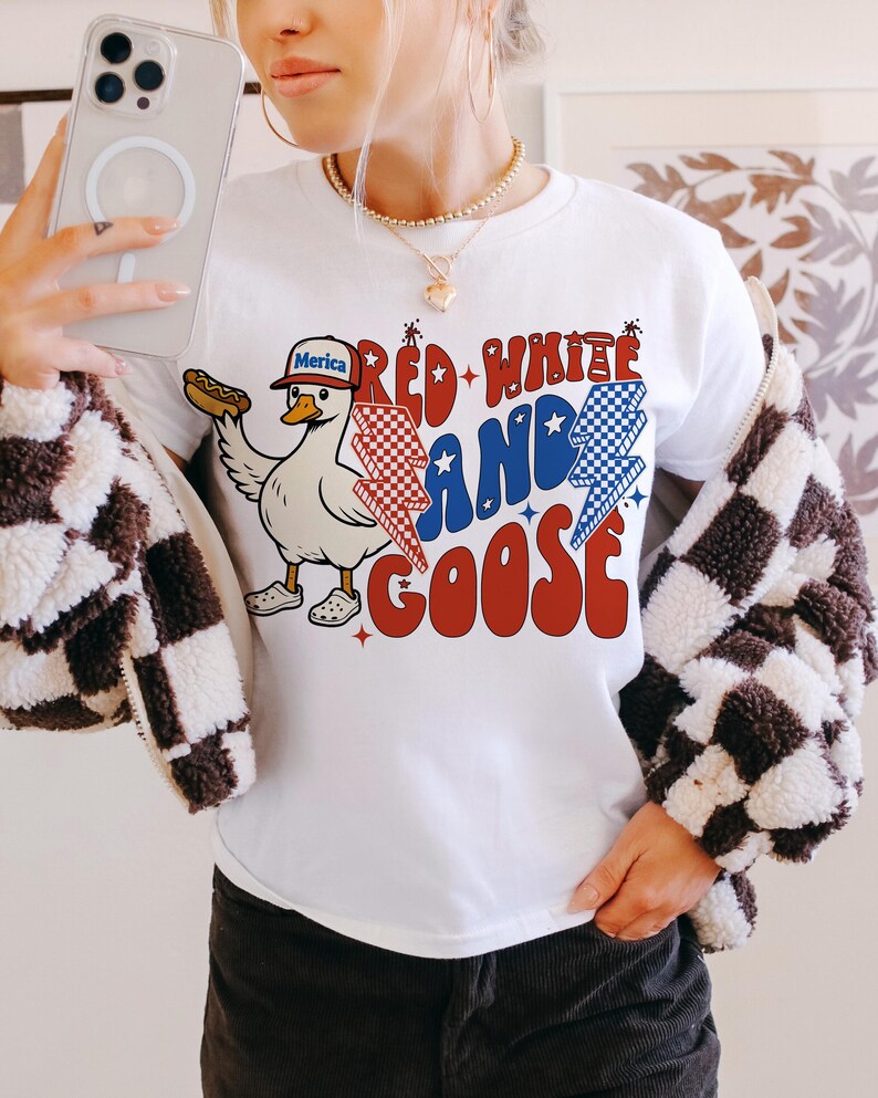 May include: White t-shirt featuring a cartoon goose wearing a baseball cap with "Merica" on it, holding a hot dog. The shirt has the text "Red White and Goose" in red, white, and blue. A casual style t-shirt.