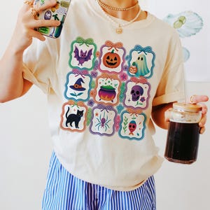 May include: Cream-colored t-shirt featuring colorful Halloween-themed illustrations. The shirt displays a bat, pumpkin, ghost, witch hat, cauldron, skull, black cat, spider, and a red-eyed skull. The person is also holding a phone and a glass of dark liquid.