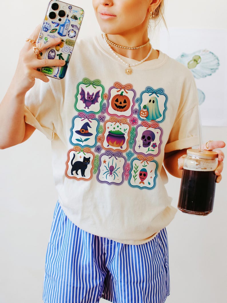 May include: Cream-colored t-shirt featuring colorful Halloween-themed illustrations. The shirt displays a bat, pumpkin, ghost, witch hat, cauldron, skull, black cat, spider, and a red-eyed skull. The person is also holding a phone and a glass of dark liquid.