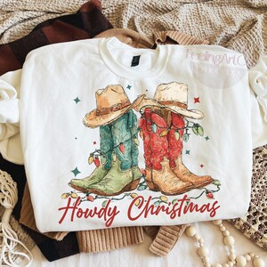 May include: White crewneck sweatshirt featuring a graphic of cowboy boots and hats decorated with Christmas lights. The boots are in shades of green, red, and brown. The text "Howdy Christmas" is written in red script below the boots.