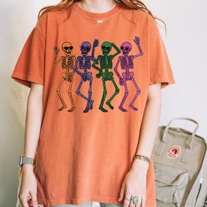 Puede incluir: Camiseta naranja con un dise&ntilde;o de cuatro esqueletos bailando. Los esqueletos son de color naranja, morado, verde y rosa. La camiseta tiene cuello redondo y mangas cortas. El dise&ntilde;o es de estilo divertido y peculiar.