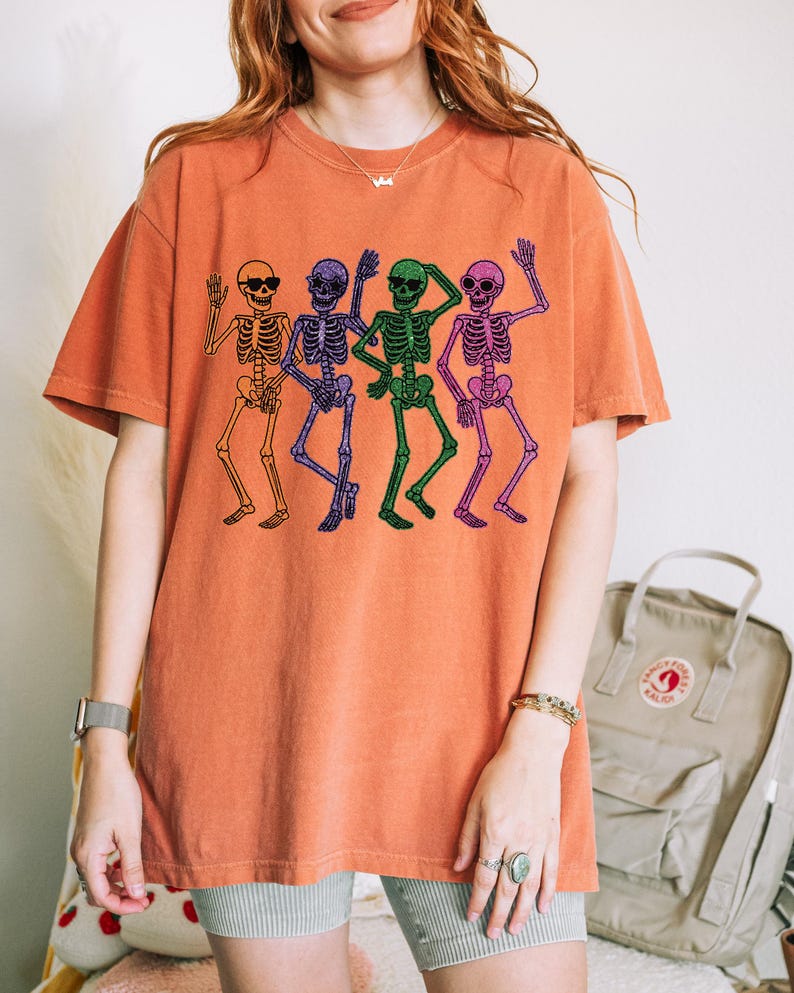 Puede incluir: Camiseta naranja con un dise&ntilde;o de cuatro esqueletos bailando. Los esqueletos son de color naranja, morado, verde y rosa. La camiseta tiene cuello redondo y mangas cortas. El dise&ntilde;o es de estilo divertido y peculiar.