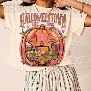 May include: A cream-colored t-shirt featuring a large orange jack-o'-lantern graphic with the text HALLOWEENTOWN EST. 1998. The pumpkin design contains a detailed illustration of a house and fence, accented by small blue and pink stars.