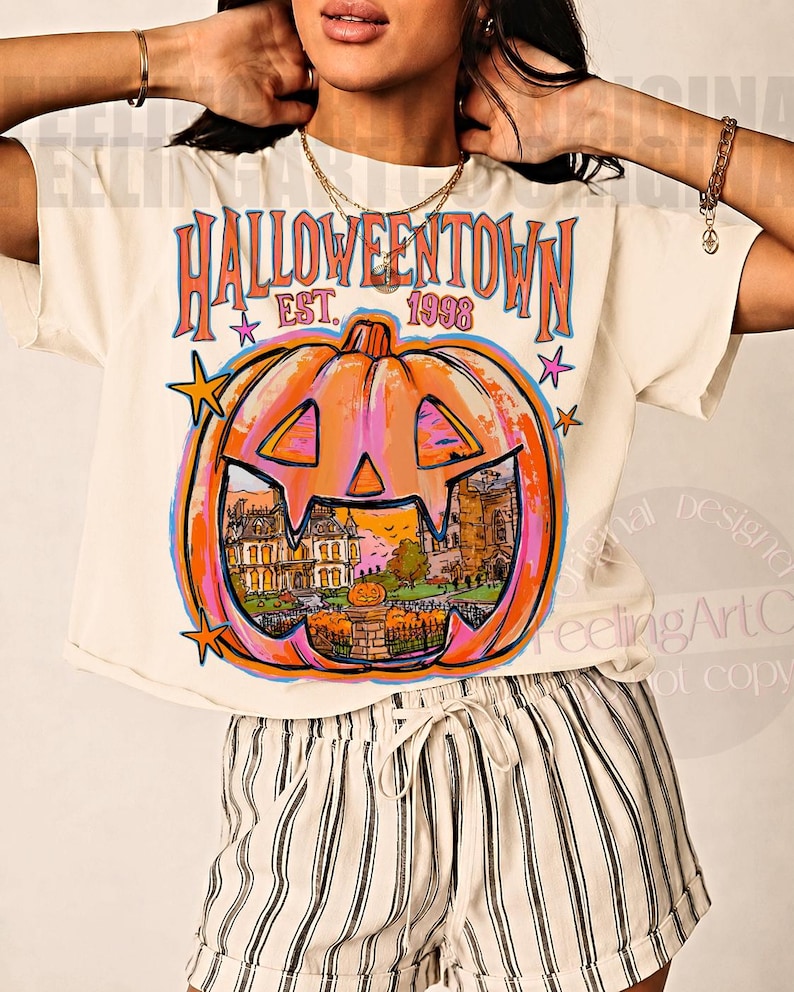 May include: A cream-colored t-shirt featuring a large orange jack-o'-lantern graphic with the text HALLOWEENTOWN EST. 1998. The pumpkin design contains a detailed illustration of a house and fence, accented by small blue and pink stars.