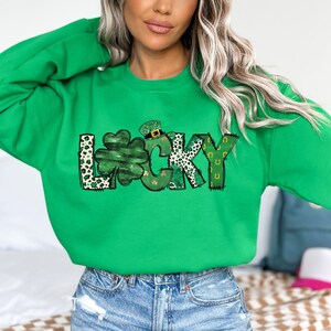 May include: A bright green sweatshirt with the word "LUCKY" in a whimsical design. The letter "L" has a leopard print pattern, the "U" is a shamrock, and the "Y" has a horseshoe design. The sweatshirt is perfect for St. Patrick's Day.