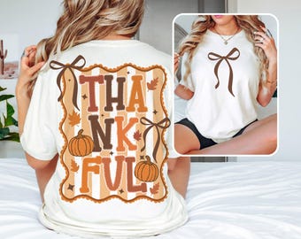 May include: Cream-colored t-shirt with a fall-themed graphic. The design features the word "THANKFUL" in brown and orange hues, pumpkins, and fall leaves. A smaller image shows a white t-shirt with a brown bow.
