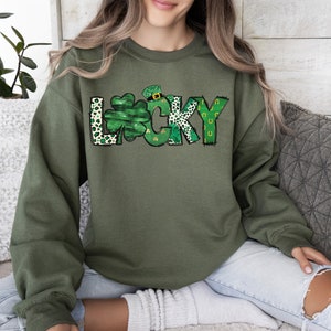 May include: Olive green sweatshirt with the word "LUCKY" in a whimsical design. The "L" features a shamrock, the "C" has a leprechaun hat, and the "K" has a leopard print pattern. The "Y" has a horseshoe design.