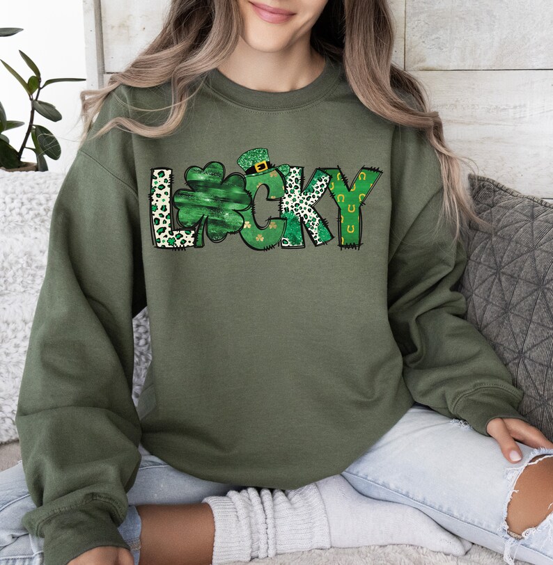 May include: Olive green sweatshirt with the word "LUCKY" in a whimsical design. The "L" features a shamrock, the "C" has a leprechaun hat, and the "K" has a leopard print pattern. The "Y" has a horseshoe design.