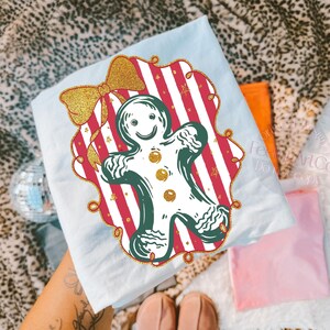 May include: A light blue t-shirt featuring a gingerbread man design. The gingerbread man is outlined in green with gold accents, set against a red and white striped background. A gold glitter bow is at the top left. The design is framed with a gold border.
