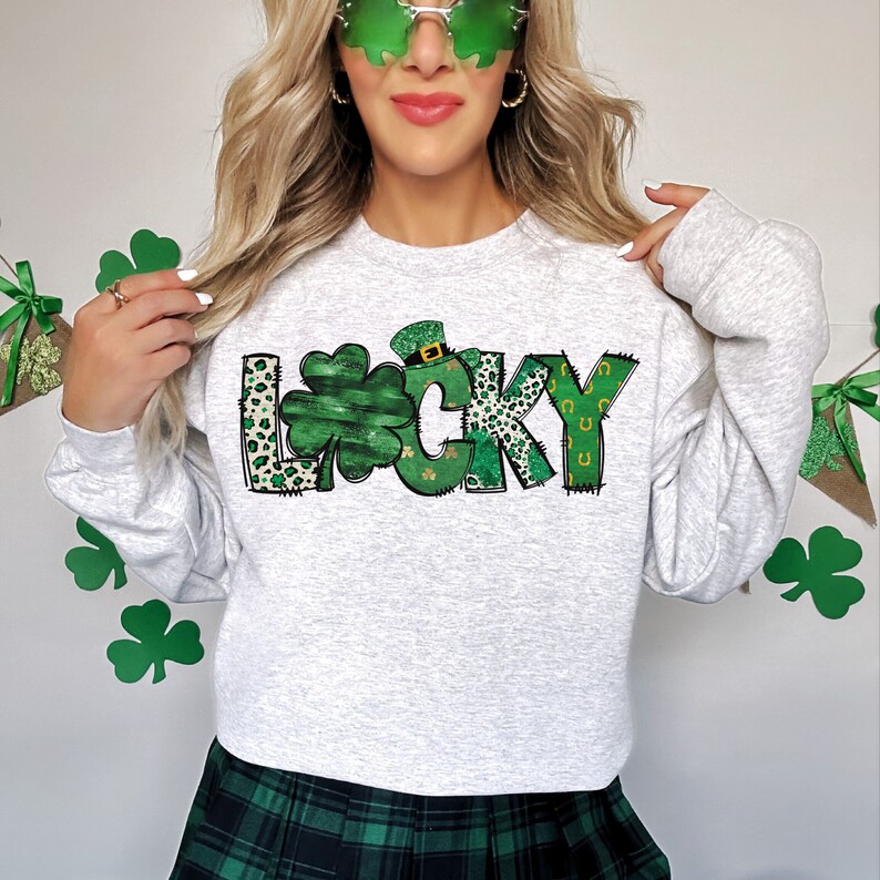 May include: A light gray sweatshirt with the word "LUCKY" in a green and black design. The "L" and "Y" have a leopard print pattern, and the "U" is a green shamrock. The "C" and "K" are green with a horseshoe design. A green shamrock and leprechaun hat are also featured.