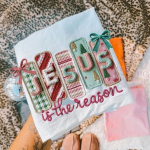 May include: White t-shirt with the embroidered text "JESUS is the reason." The word "JESUS" is spelled out with colorful, decorative rectangles. Each rectangle has a different pattern and color, including green plaid, red stripes, and pink with Christmas trees.