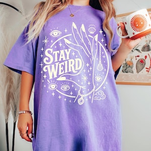 May include: A purple t-shirt with the words "Stay Weird" in a circular design. The design includes a hand, eyes, stars, and a seashell. The shirt is a short-sleeved, oversized style.