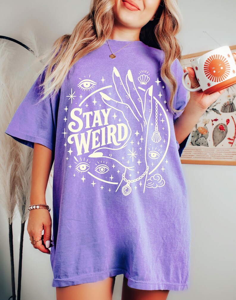 May include: A purple t-shirt with the words "Stay Weird" in a circular design. The design includes a hand, eyes, stars, and a seashell. The shirt is a short-sleeved, oversized style.