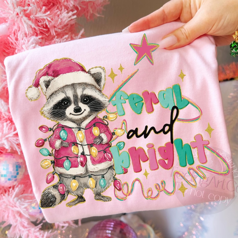 May include: A light pink t-shirt featuring a cartoon raccoon wearing a Santa hat and a pink coat adorned with Christmas lights. The text "feral and bright" is printed on the shirt with a star and other decorative elements.
