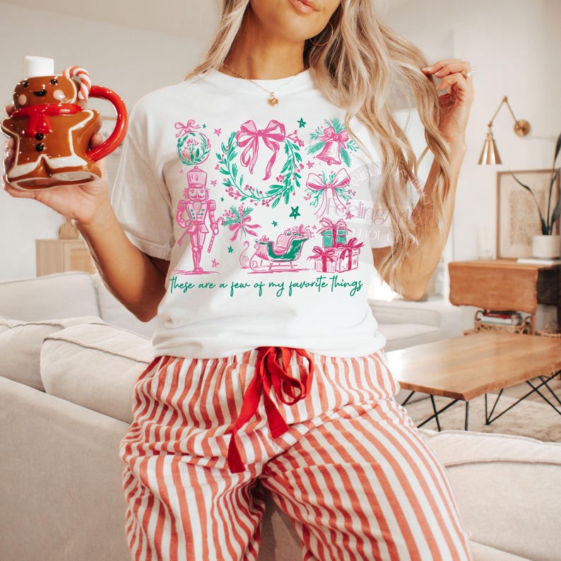 May include: White t-shirt with a pink and green Christmas graphic and the text "These are a few of my favorite things." The shirt is paired with red and white striped pajama pants. A gingerbread man mug is held in the left hand.