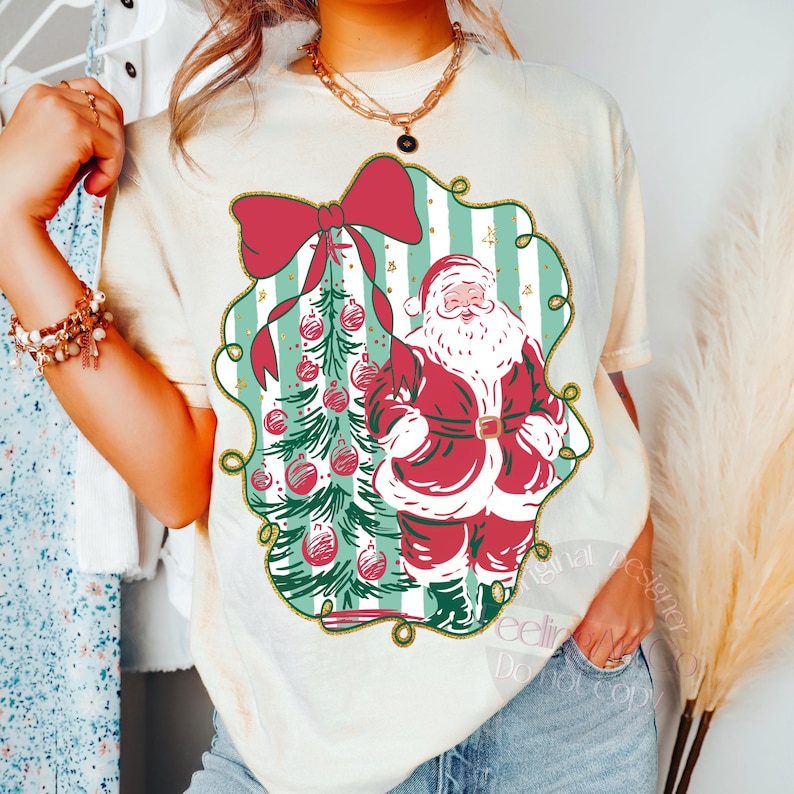 May include: Cream-colored t-shirt featuring a Christmas design. The design includes a red bow, a Christmas tree with ornaments, and Santa Claus in a red suit. The design is framed by a green and gold border.