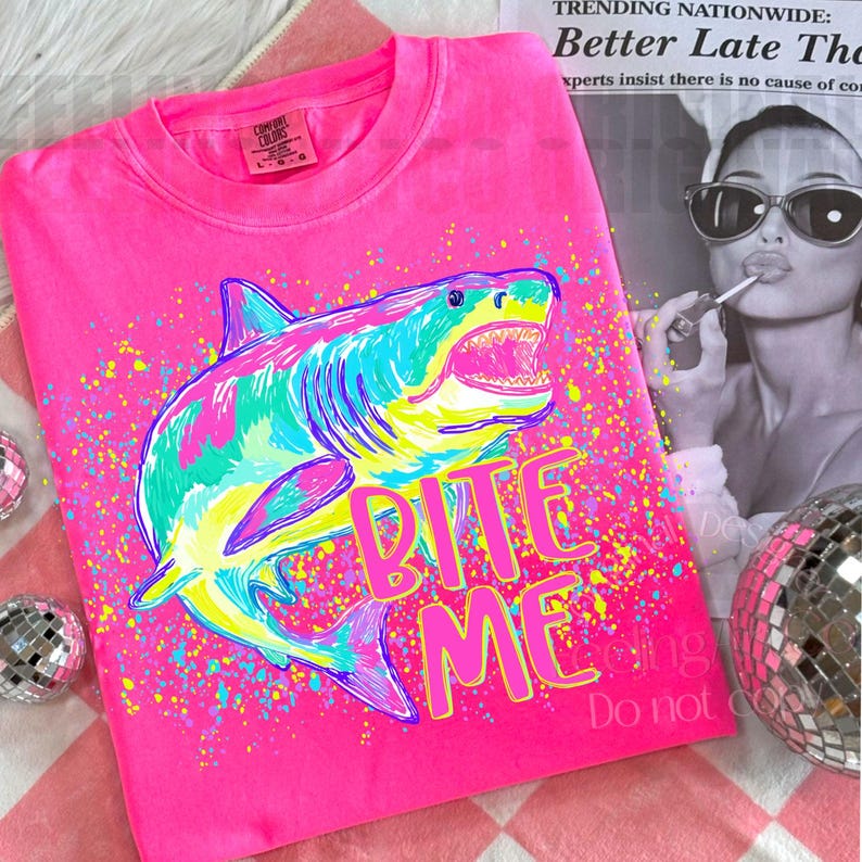 Op de afbeelding: Felroze T-shirt met een kleurrijke haai-afbeelding en de tekst "Bite Me". De haai is afgebeeld in tinten blauw, groen en geel. Het T-shirt heeft een ronde hals en korte mouwen.