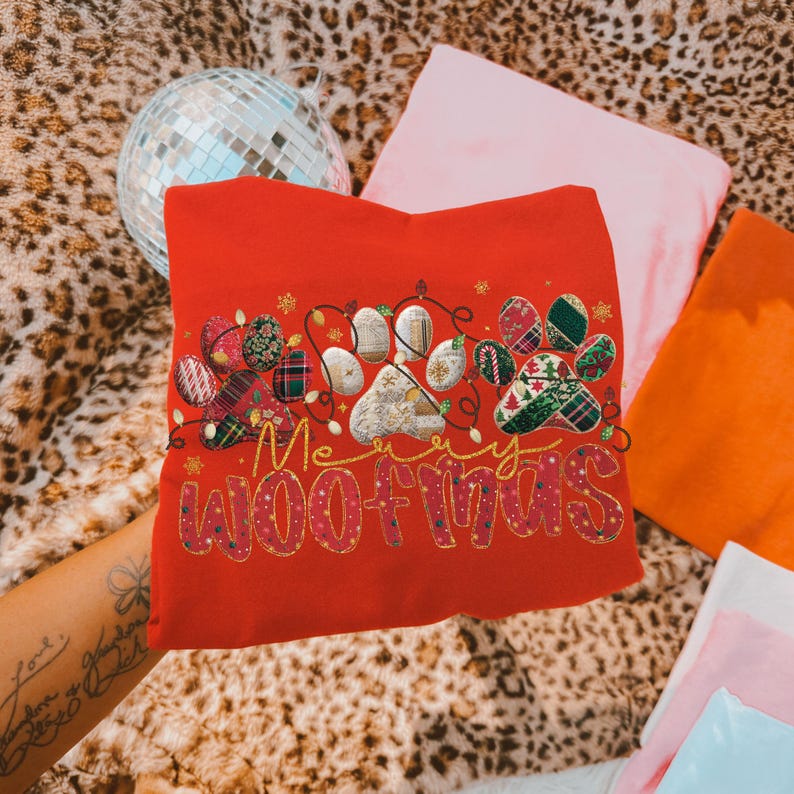 Puede incluir: Una sudadera roja con el texto dorado "Merry Woofmas". El dise&ntilde;o incluye una huella de pata y adornos decorativos. Un art&iacute;culo festivo para la temporada navide&ntilde;a.