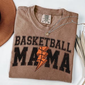 May include: A brown t-shirt with the text "Basketball Mama" in black with a lightning bolt graphic in orange and brown. The shirt is made of a soft, comfortable material.