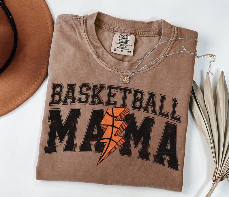 May include: A brown t-shirt with the text "Basketball Mama" in black with a lightning bolt graphic in orange and brown. The shirt is made of a soft, comfortable material.