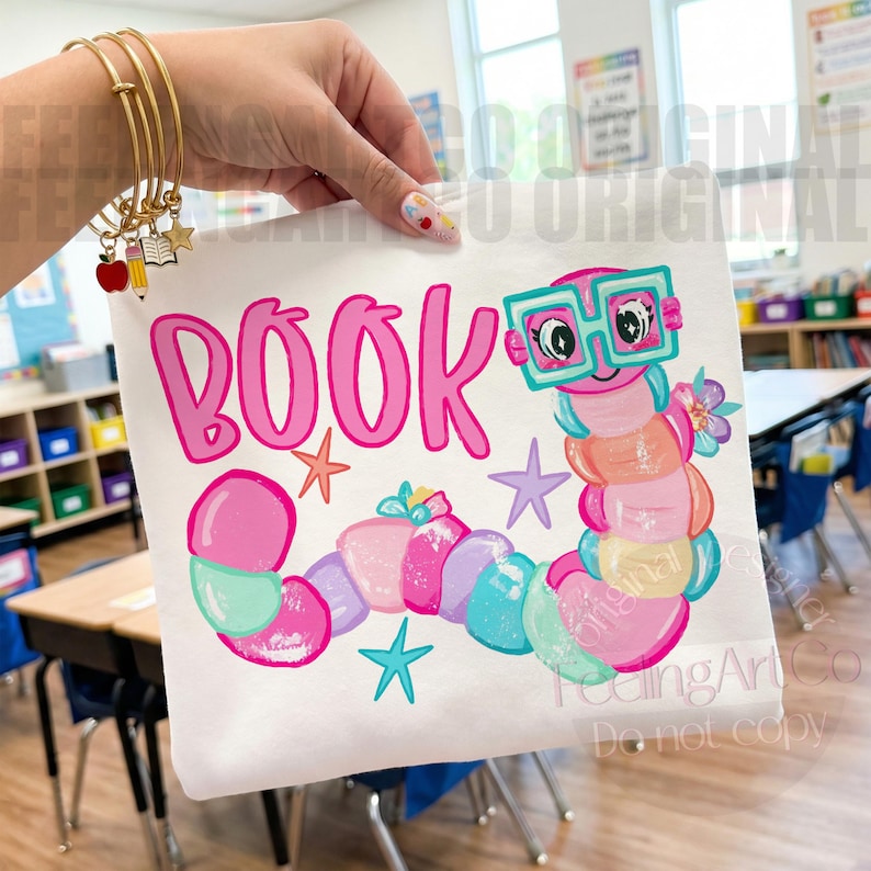 May include: A white t-shirt featuring a colorful cartoon bookworm wearing teal glasses, surrounded by the pink text "BOOK" and small star shapes. The design includes a multi-colored segmented worm body with floral accents and a bright pink font.