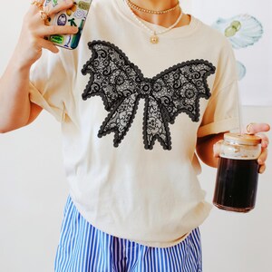 May include: Cream-colored t-shirt featuring a large black lace bow design. The bow has intricate details and a scalloped edge. The model is wearing blue and white striped shorts and holding a phone with a colorful case.