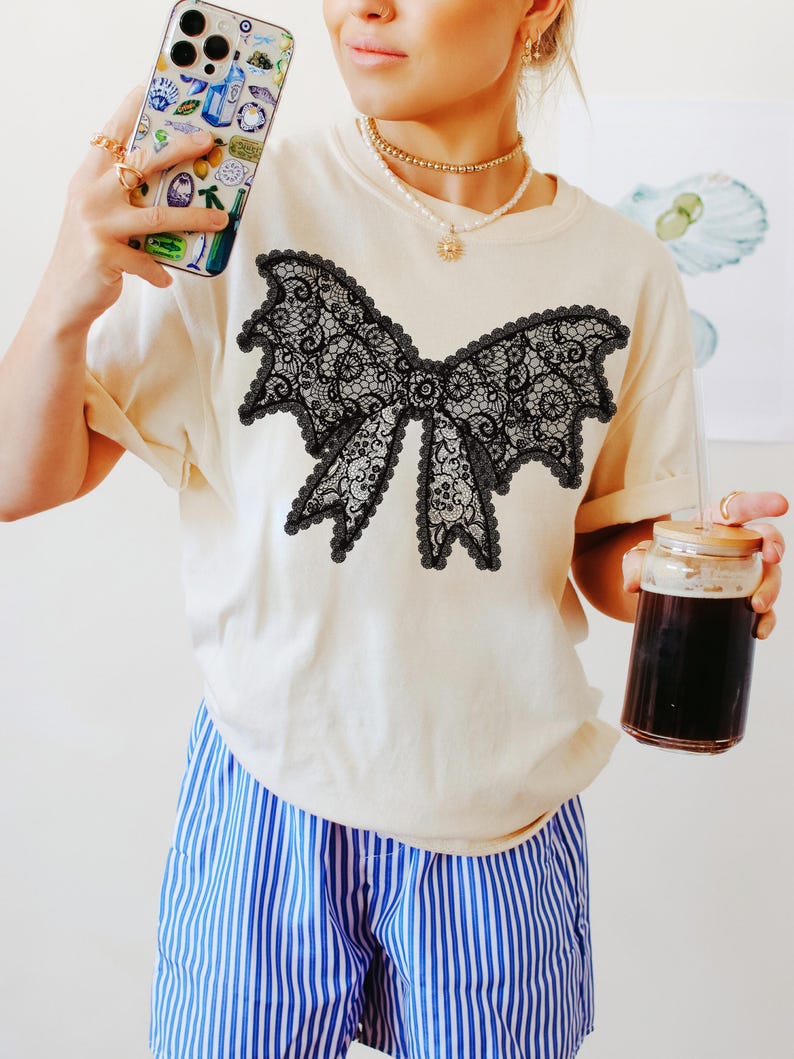 May include: Cream-colored t-shirt featuring a large black lace bow design. The bow has intricate details and a scalloped edge. The model is wearing blue and white striped shorts and holding a phone with a colorful case.