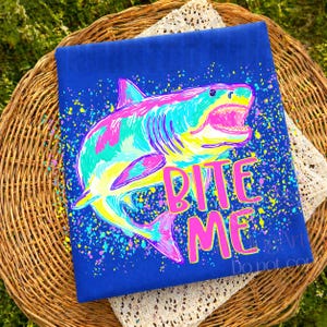 Op de afbeelding: Een blauw t-shirt met een kleurrijke haaienillustratie en de tekst "BITE ME" in roze. De haai is afgebeeld in tinten roze, turquoise, geel en blauw, met een spetterverfeffect.