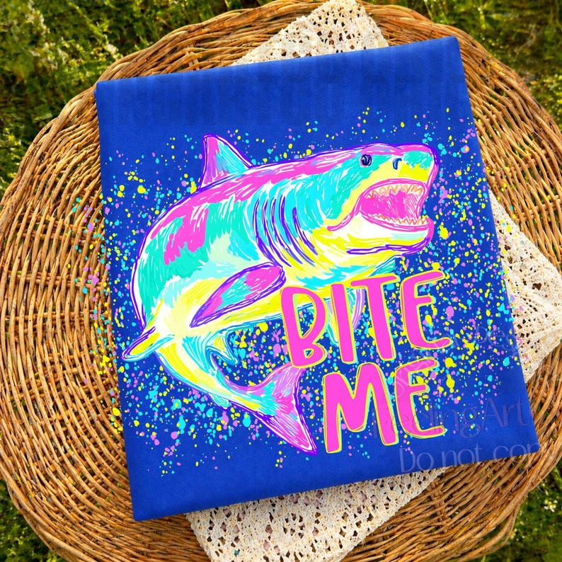 Op de afbeelding: Een blauw t-shirt met een kleurrijke haaienillustratie en de tekst "BITE ME" in roze. De haai is afgebeeld in tinten roze, turquoise, geel en blauw, met een spetterverfeffect.